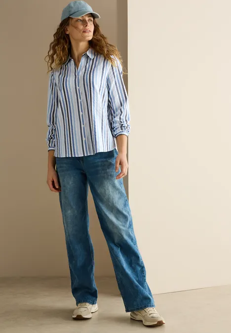 Frau mit Jeans, gestreiftem Hemd und Kappe steht vor heller Wand.